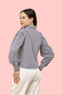 A woman wearing a black and white gingham long-sleeve blouse with puffed balloon sleeves, paired with cream-colored pants, posing against a solid light pink background.