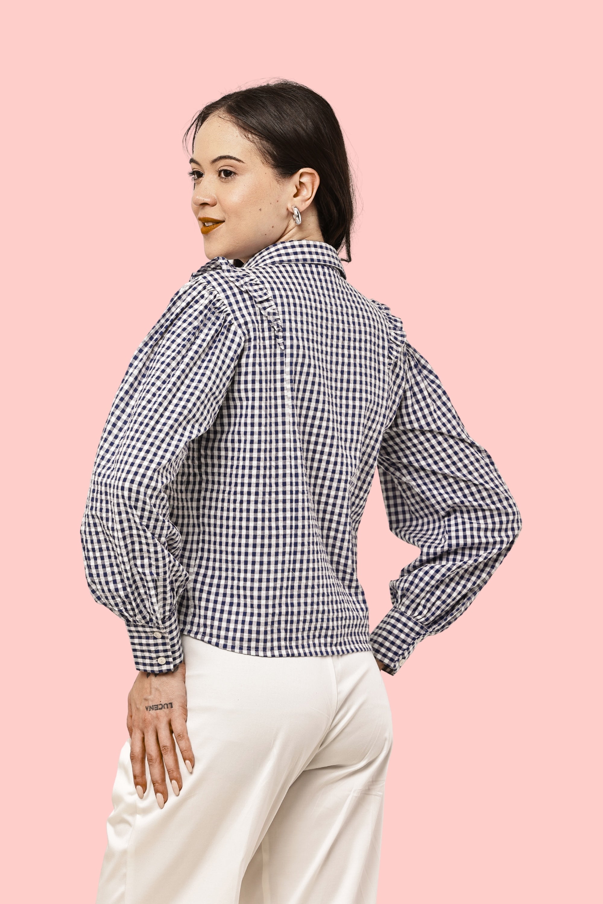 A woman wearing a black and white gingham long-sleeve blouse with puffed balloon sleeves, paired with cream-colored pants, posing against a solid light pink background.