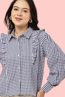 A woman wearing a navy and white gingham long-sleeve blouse with ruffle details on the shoulders and chest, set against a solid pink background.