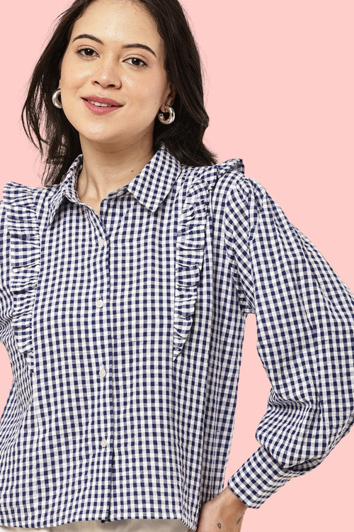 A woman wearing a navy and white gingham long-sleeve blouse with ruffle details on the shoulders and chest, set against a solid pink background.