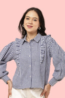 A woman wearing a navy and white gingham long-sleeve blouse with ruffle details on the shoulders and chest, paired with white pants, set against a solid pink background.