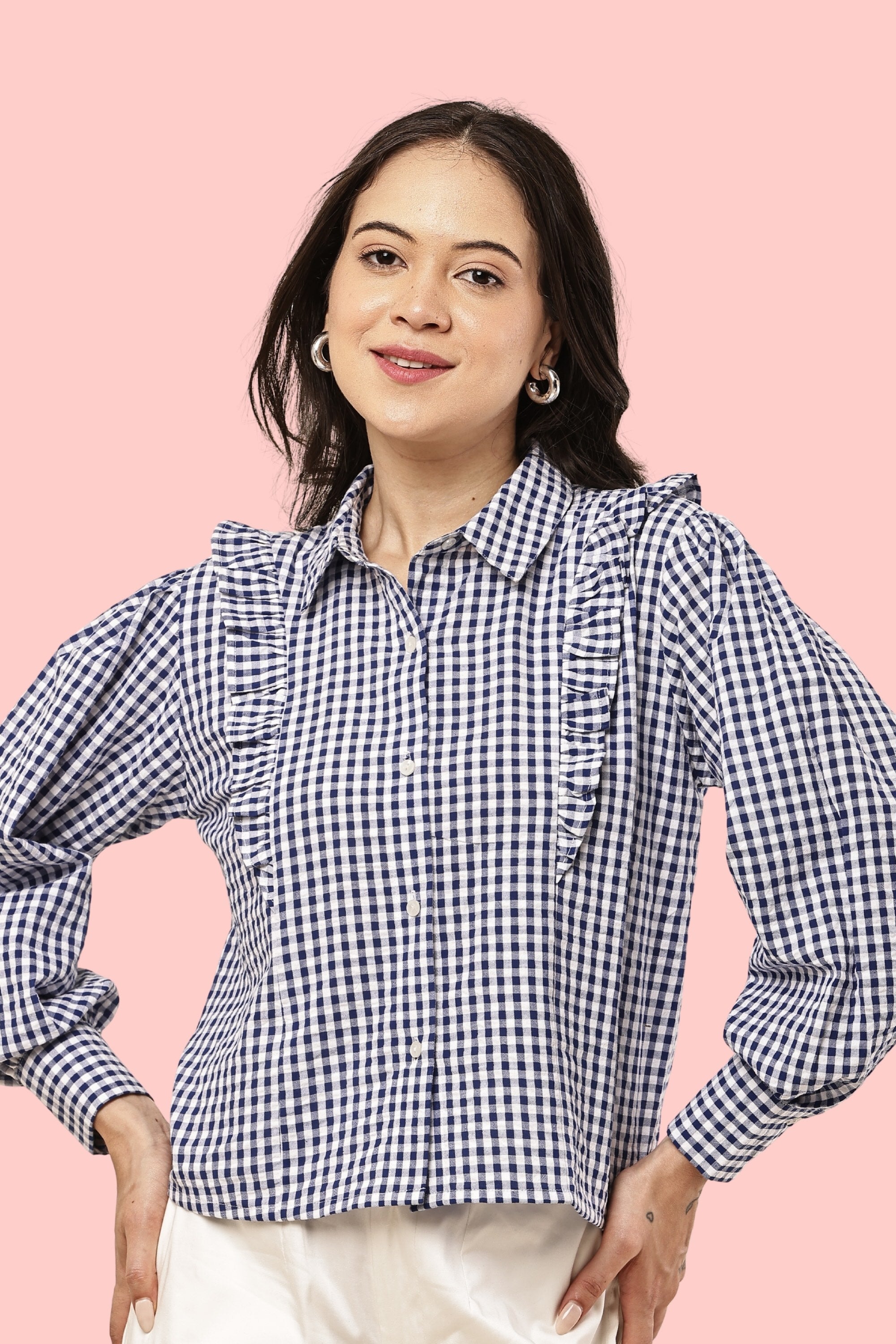 A woman wearing a navy and white gingham long-sleeve blouse with ruffle details on the shoulders and chest, paired with white pants, set against a solid pink background.