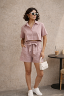 Woman wearing a pink checkered outfit with sunglasses and a white handbag against a beige wall.