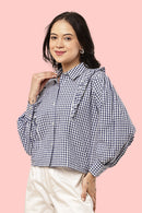 A woman wearing a navy and white gingham check long-sleeve blouse with ruffle details on the shoulders and collar, paired with cream-colored pants, set against a solid pink background.