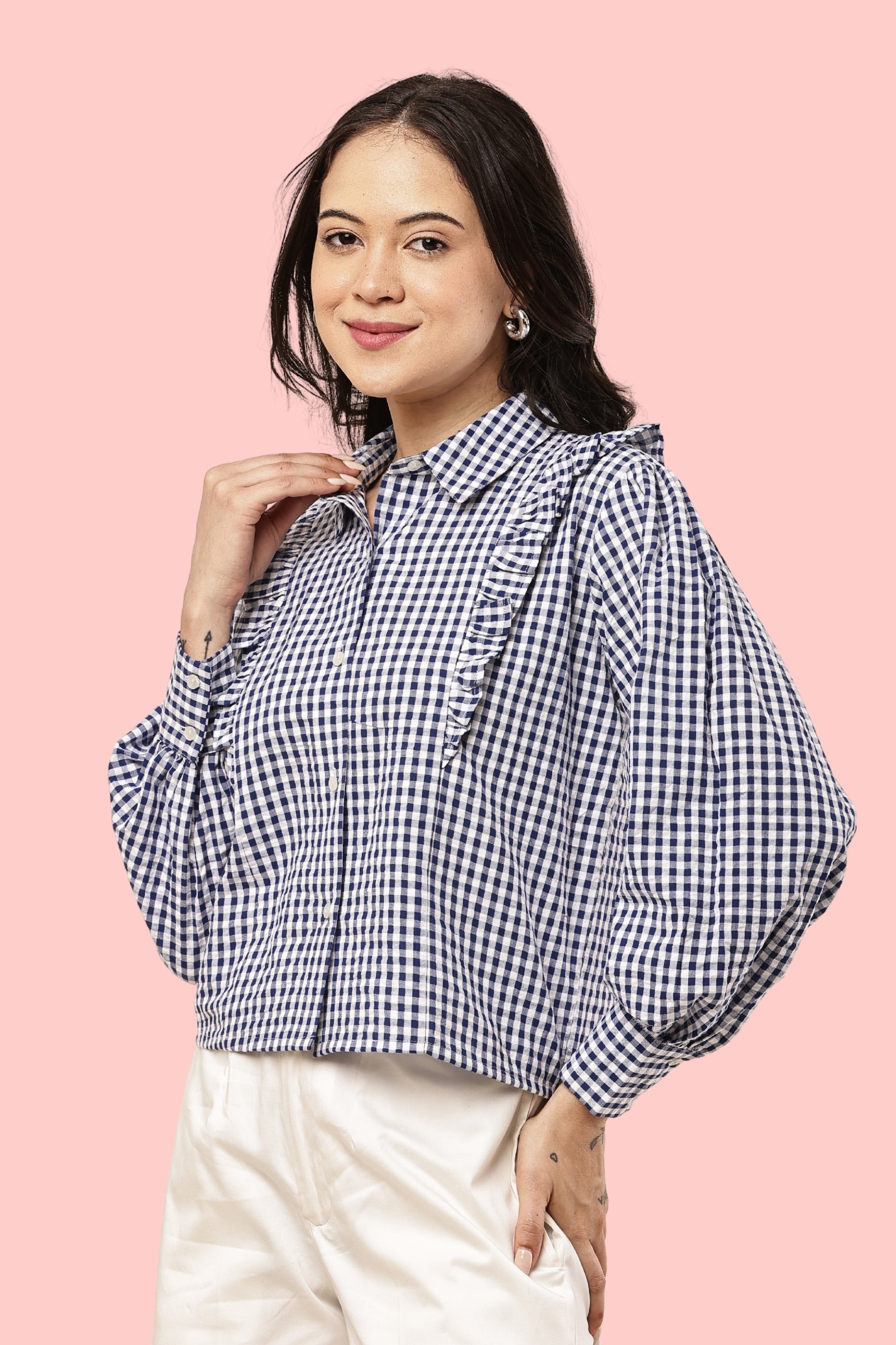 A woman wearing a navy and white gingham check long-sleeve blouse with ruffle details on the shoulders and collar, paired with cream-colored pants, set against a solid pink background.