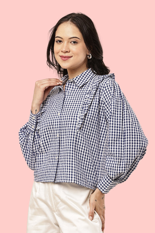 A woman wearing a navy and white gingham check long-sleeve blouse with ruffle details on the shoulders and collar, paired with cream-colored pants, set against a solid pink background.