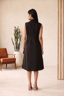 A woman viewed from behind wearing a sleeveless, collared black dress with a Tucks waist, standing in a minimalist beige interior setting.