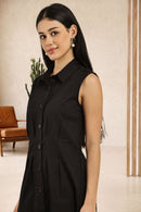 A woman with long black hair is wearing a sleeveless black button-front collared dress, photographed indoors against a light beige wall with a brown chair and plant in the background.