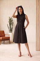 A woman models a sleeveless, button-front midi dress in a warm Black color. The dress has a collared neckline and a flared skirt, shown in a styled indoor setting with a beige textured wall, arches, and a small black handbag.
