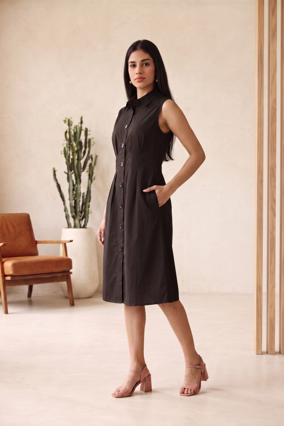 A woman modeling a sleeveless black midi shirt dress with a button-down front and side pockets, standing in a minimalist beige room with a cactus and a brown chair in the background.
