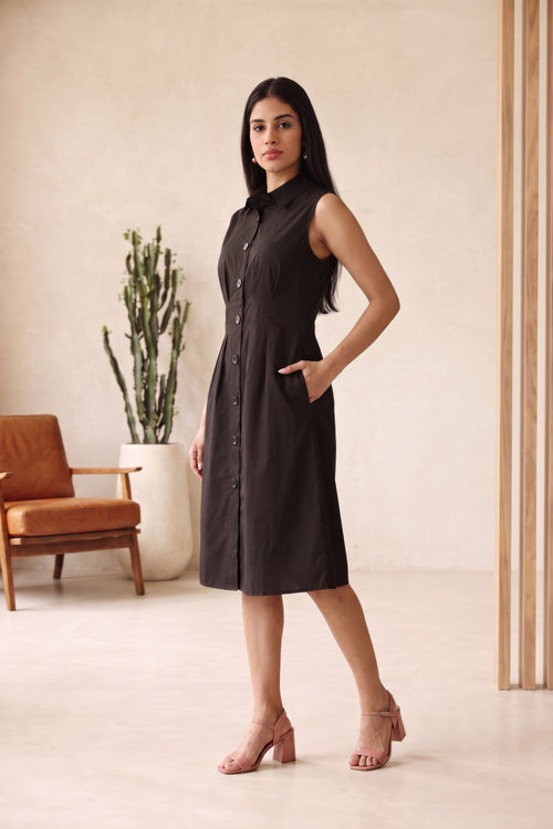 A woman modeling a sleeveless black midi shirt dress with a button-down front and side pockets, standing in a minimalist beige room with a cactus and a brown chair in the background.