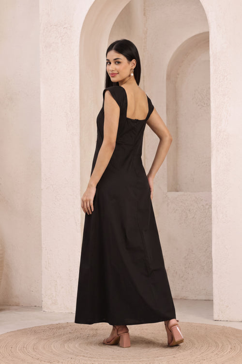 A woman modeling a sleeveless black maxi dress with a draped open back, standing in a beige stone archway setting.