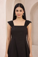 A woman with long black hair wearing a black sleeveless dress with a square neckline and ruffled cap sleeves, standing against a light beige textured wall with arches.