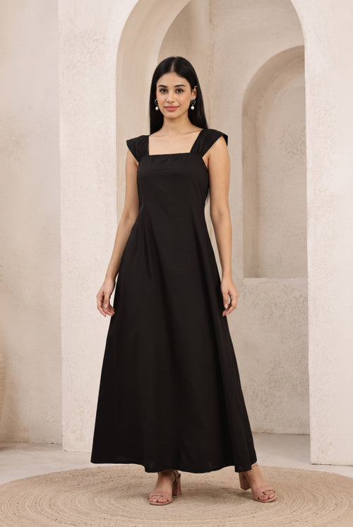 A woman modeling a black sleeveless maxi dress with a square neckline and flared skirt, standing in front of a beige stone archway background.