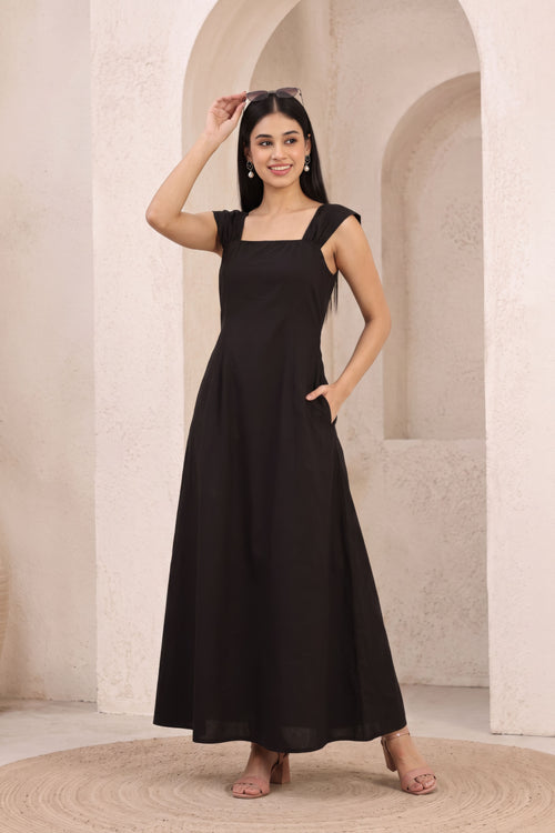 A woman modeling a black sleeveless maxi dress with a square neckline and side pockets, standing in front of a beige stone archway background.