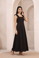 A woman modeling a black sleeveless maxi dress with a square neckline and a flowing skirt, standing in front of a beige stone archway background.