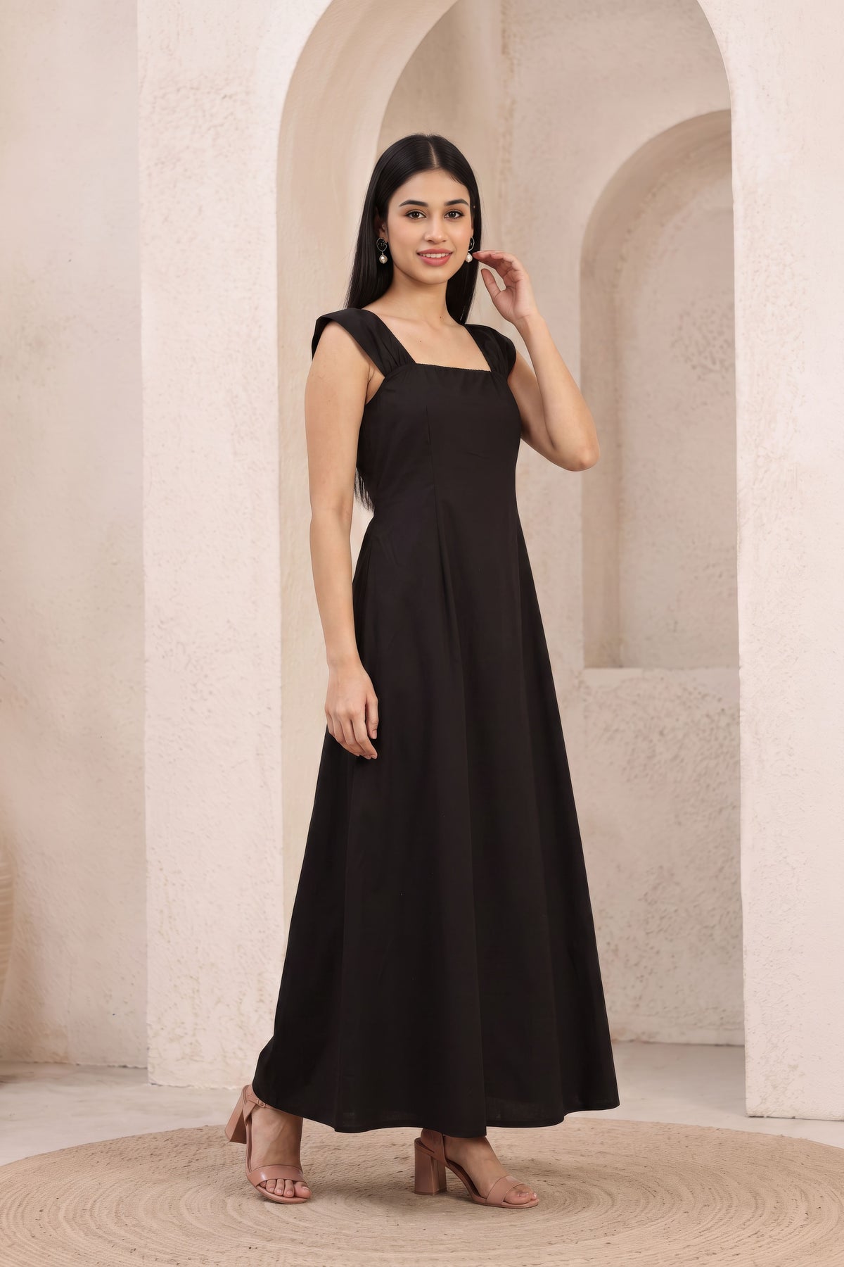 A woman modeling a black sleeveless maxi dress with a square neckline and a flowing skirt, standing in front of a beige stone archway background.