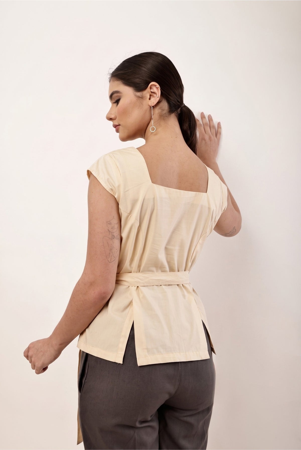 A woman shown from the back wearing a light beige wrap-style top with a square neckline and tied waist, paired with dark gray pants, against a plain off-white background.