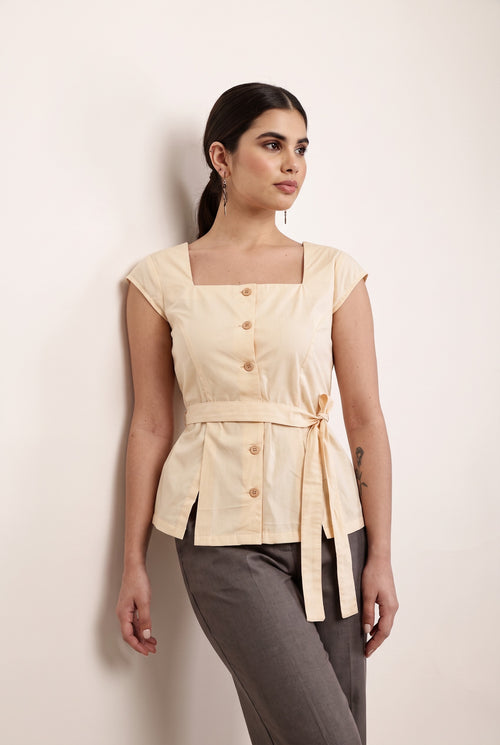 A woman wearing a peach-colored sleeveless top with a square neckline, button-front detail, and a tied waist sash, paired with dark pants and blue-tinted sunglasses, against a plain light beige background.