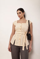 A woman modeling a terracotta brown short-sleeve top with a square neckline, front buttons, and a matching waist tie, paired with black pants, standing against a plain off-white wall.