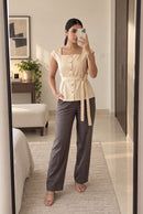 A woman takes a mirror selfie in a bedroom setting, wearing a beige peplum-style top with a square neckline, short cap sleeves, and a matching waist tie, paired with dark gray wide-leg trousers.