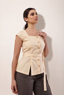 A woman modeling a light beige peplum top with a square neckline, cap sleeves, front buttons, a matching tie belt, and dark gray trousers, set against a plain off-white background.