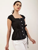 A woman modeling a black short-sleeve peplum top with a square neckline, front button closure, and side tie detail, paired with white pants against a plain light background.
