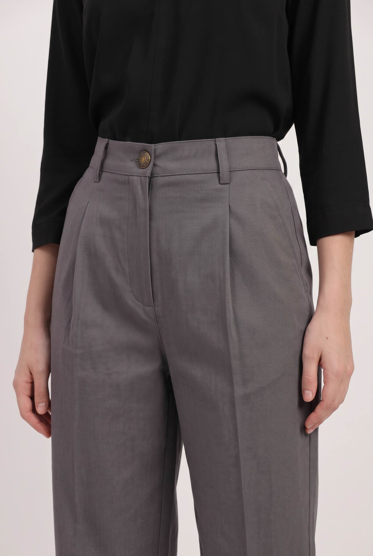 A close-up of a person wearing high-waisted gray pleated trousers with a black long-sleeve top, shown from the chest down against a plain light background.