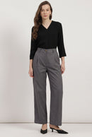 A woman modeling a black long-sleeve V-neck top and high-waisted gray wide-leg trousers with black pointed-toe heels, standing against a plain light gray studio background.