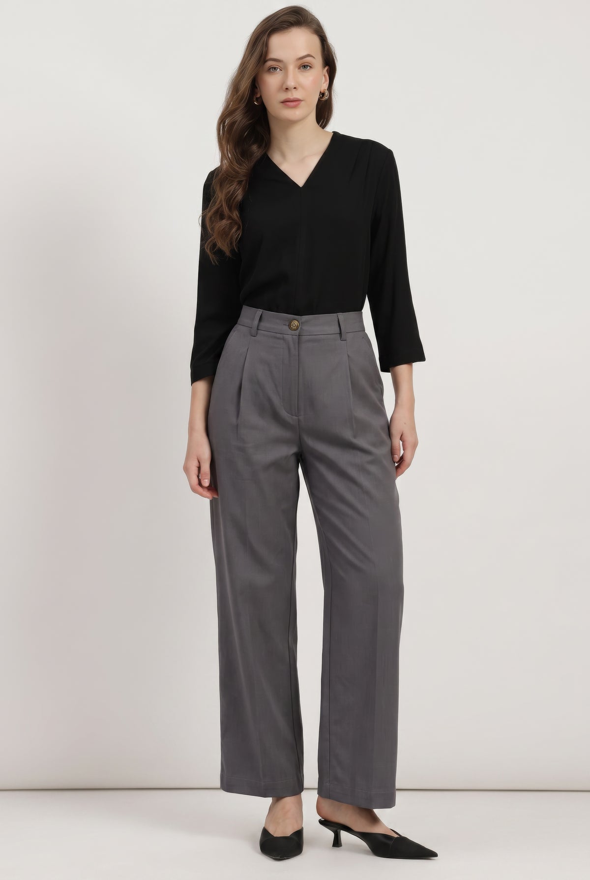 A woman modeling a black long-sleeve V-neck top and high-waisted gray wide-leg trousers with black pointed-toe heels, standing against a plain light gray studio background.