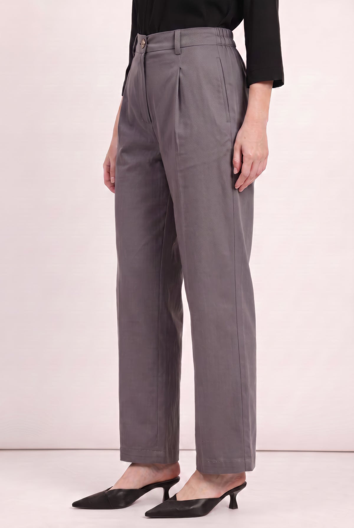 A model wearing high-waisted, wide-leg gray trousers with a front pleat detail, styled with a black top and black pointed-toe mule heels against a light pink studio background.