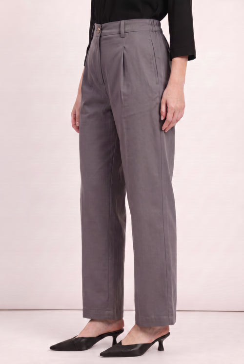 A model wearing high-waisted, wide-leg gray trousers with a front pleat detail, styled with a black top and black pointed-toe mule heels against a light pink studio background.