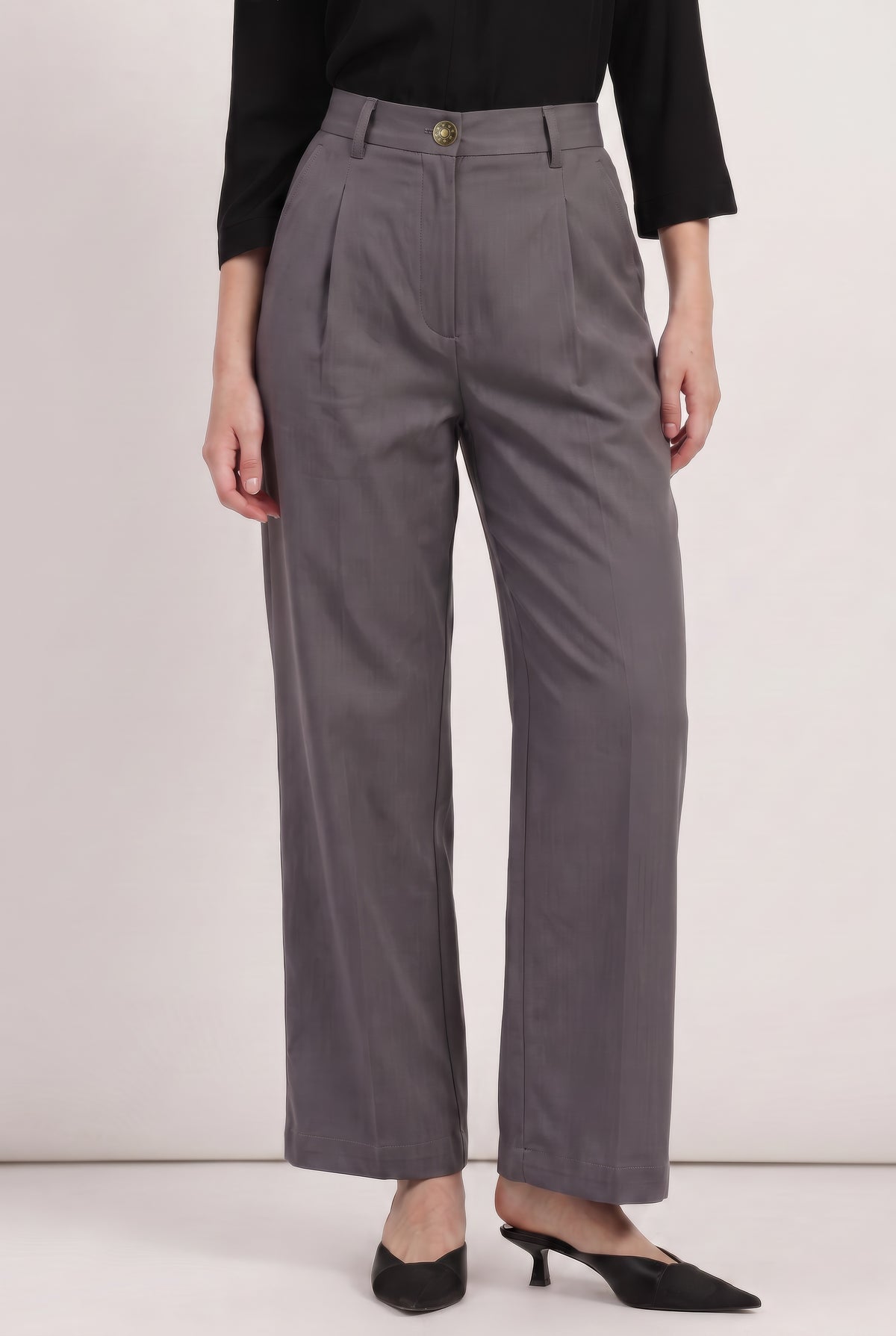 A model is wearing high-waisted, wide-leg gray trousers with pleats, styled with a black long-sleeve top and black pointed-toe mule heels against a plain off-white studio background.