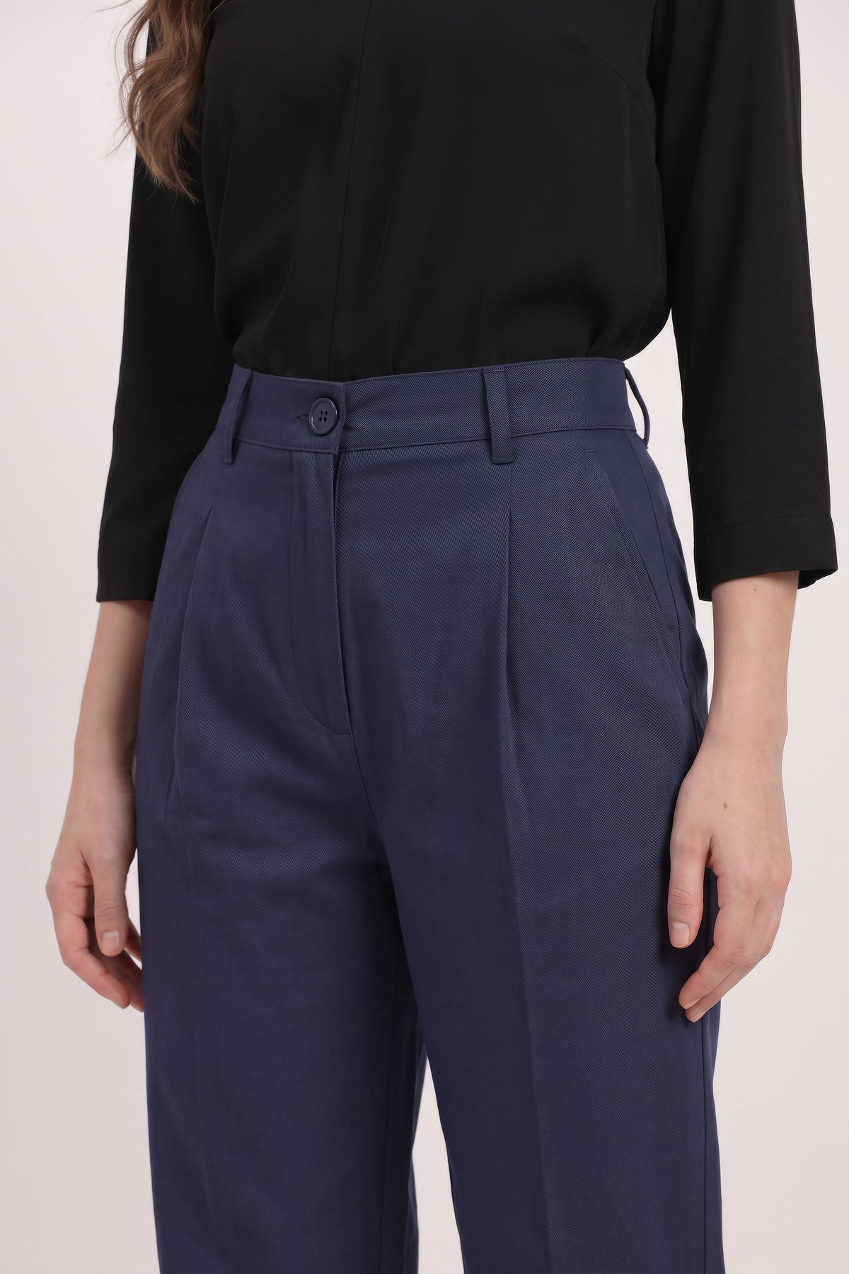 A close-up of a person wearing navy blue high-waisted pleated trousers and a black long-sleeve top, shown from the chest down against a plain light background.
