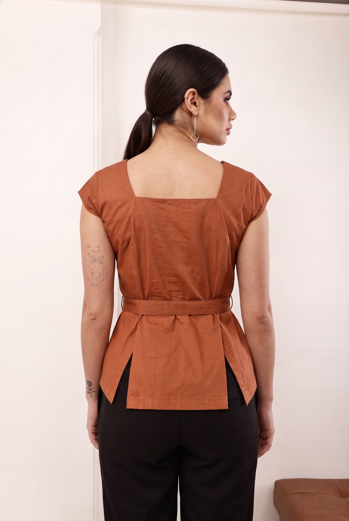 A woman shown from the back wearing a rust-orange peplum top with a square neckline and a matching waist tie, paired with black pants, in an indoor setting.