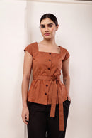 A woman modeling a terracotta brown short-sleeve top with a square neckline, front buttons, and a matching waist tie, paired with black pants, standing against a plain off-white wall.