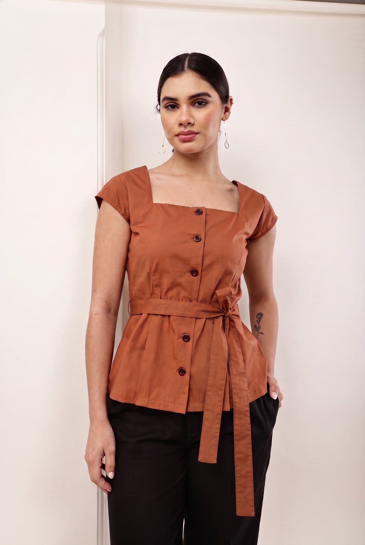 A woman modeling a terracotta brown short-sleeve top with a square neckline, front buttons, and a matching waist tie, paired with black pants, standing against a plain off-white wall.