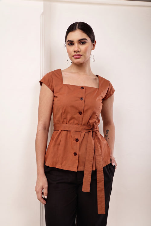A woman modeling a terracotta brown short-sleeve top with a square neckline, front buttons, and a matching waist tie, paired with black pants, standing against a plain off-white wall.