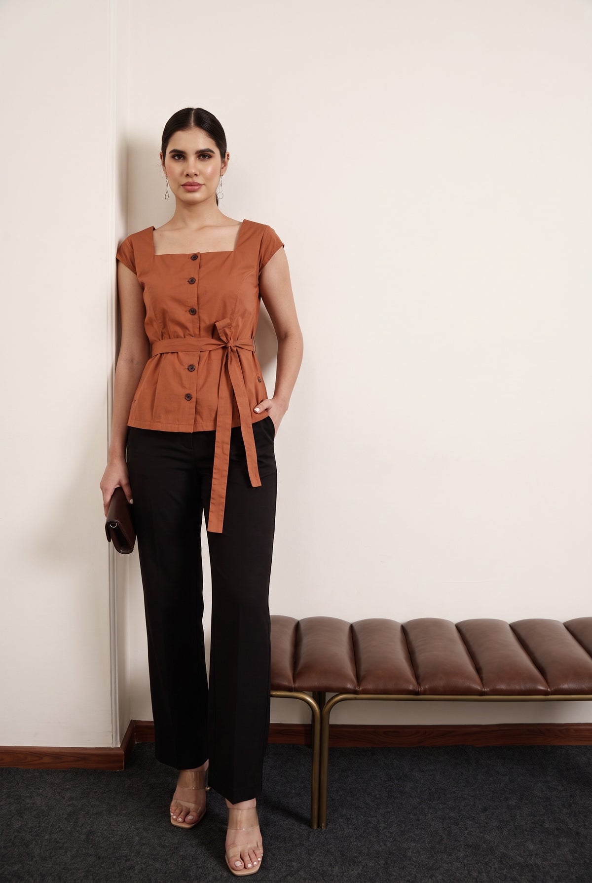 A woman stands indoors wearing a rust-orange belted top with cap sleeves and black wide-leg pants, holding a small dark clutch. A brown bench is against a plain light wall behind her.