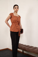 A woman modeling a rust-orange short-sleeve button-front top with a tie waist, paired with black trousers, standing in a minimalist room with a brown bench against a white wall.
