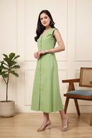 A woman modeling a sleeveless light green midi dress with a buttoned front, standing in a styled room with a potted plant and a wooden chair.