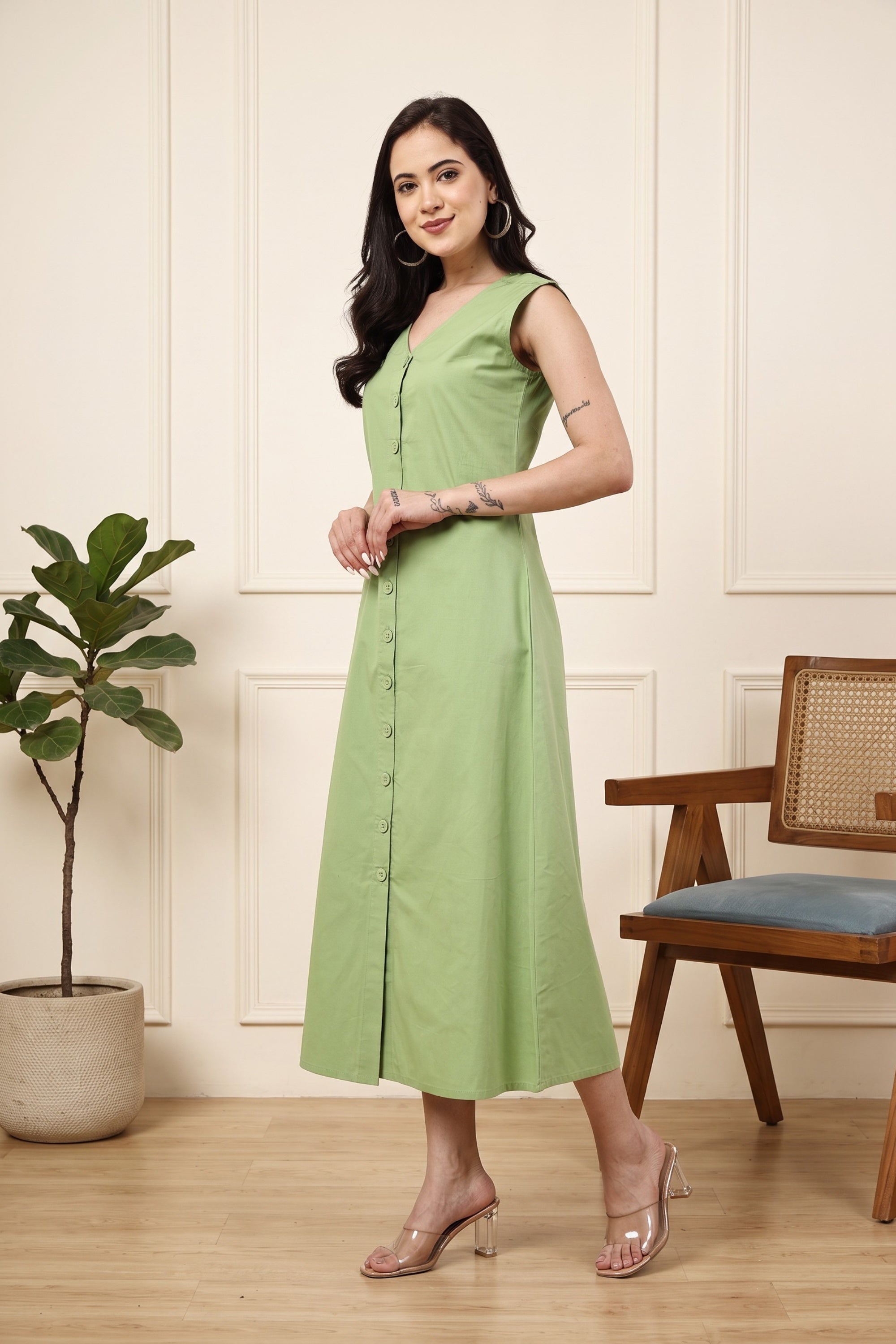 A woman modeling a sleeveless light green midi dress with a buttoned front, standing in a styled room with a potted plant and a wooden chair.