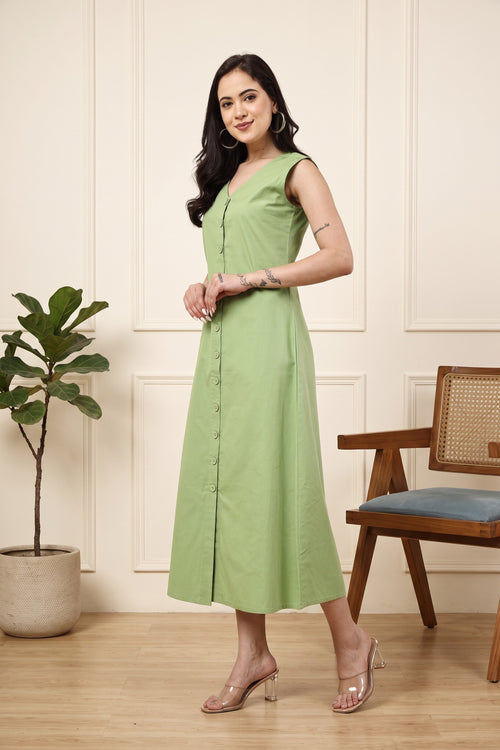 A woman modeling a sleeveless light green midi dress with a buttoned front, standing in a styled room with a potted plant and a wooden chair.