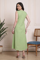 A woman wearing a sleeveless light green midi dress, photographed from the back in an indoor room with a cream paneled wall, a potted plant, and a wooden chair with a blue seat.