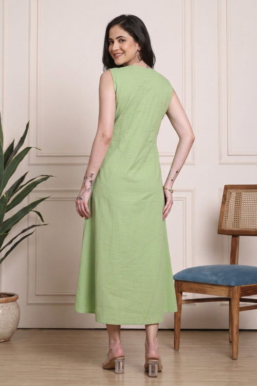 A woman wearing a sleeveless light green midi dress, photographed from the back in an indoor room with a cream paneled wall, a potted plant, and a wooden chair with a blue seat.