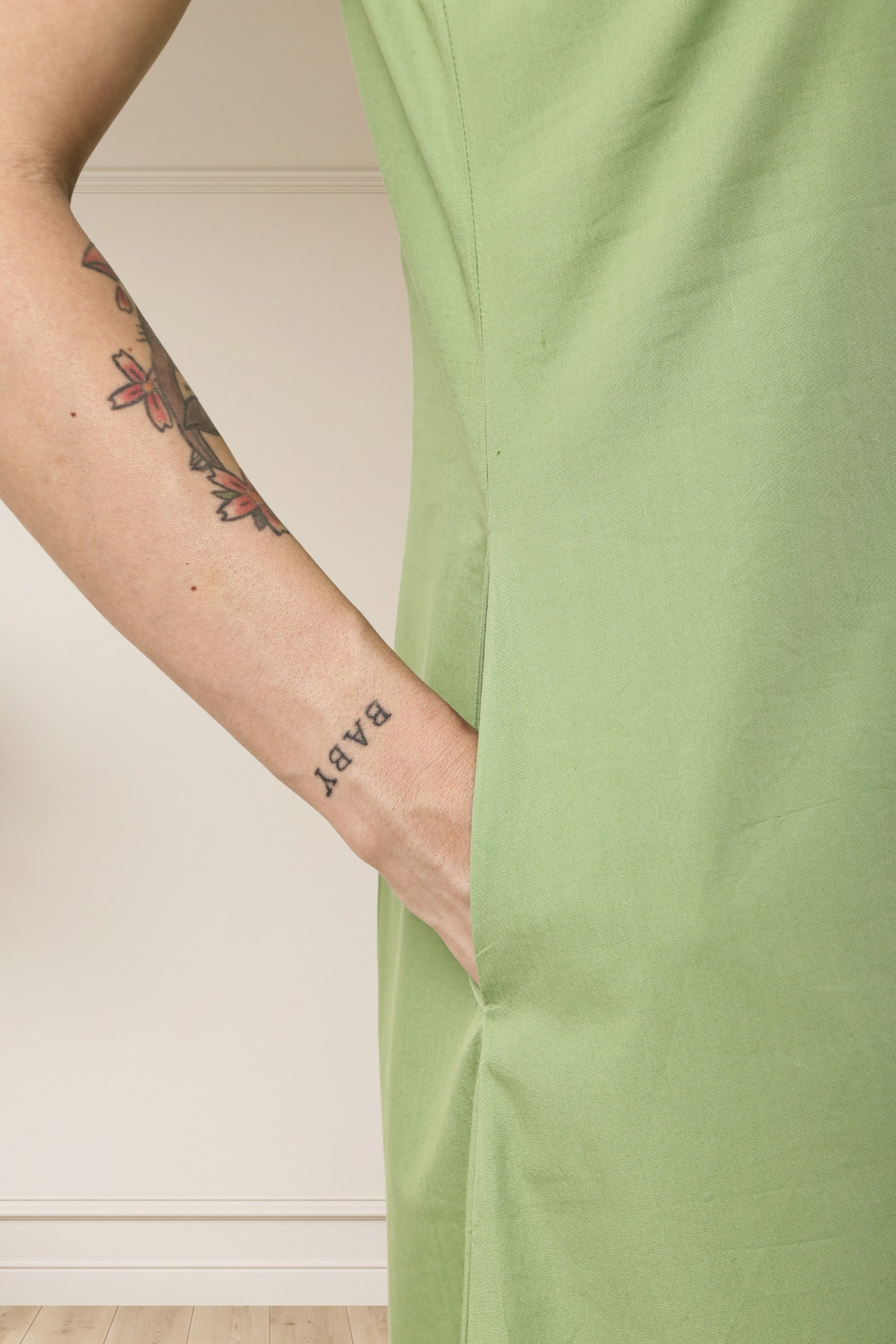 Close-up of a person wearing a light sage green dress, showing the side pocket and hand with a 'BABY' tattoo, against a neutral indoor background.