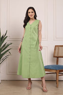 A woman modeling a sleeveless, light green midi dress with a button-down front, standing in a room with a cream paneled wall, a wooden chair with a blue cushion, and a potted plant.
