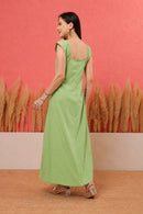 A woman wearing a light green sleeveless maxi dress with a square back, standing in front of a textured coral-pink wall with pampas grass decor.