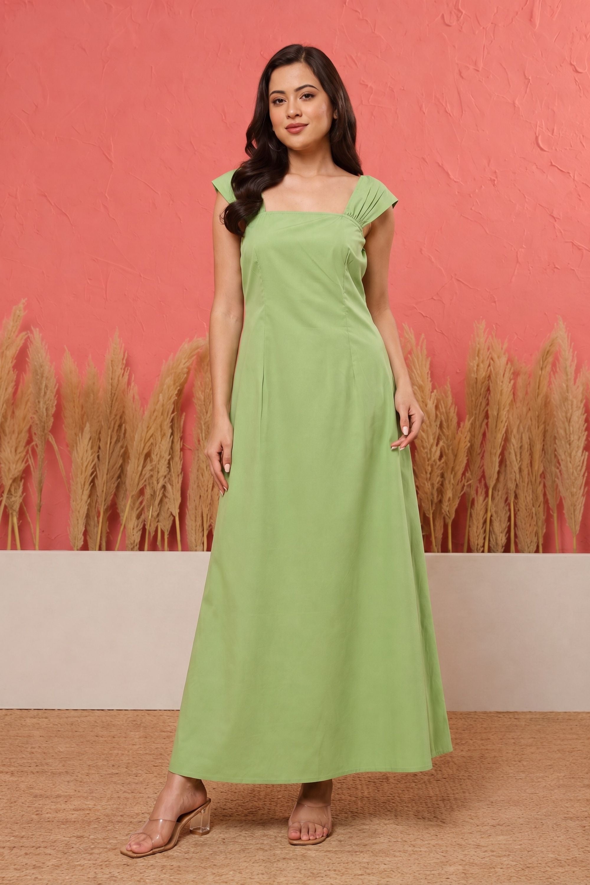 A woman modeling a light green maxi dress with a square neckline and cap sleeves, standing against a textured coral-pink wall with pampas grass decor.