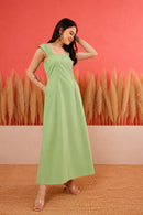 A woman modeling a light green sleeveless maxi dress with a square neckline and side pockets, standing against a coral-pink wall with dried pampas grass decor.